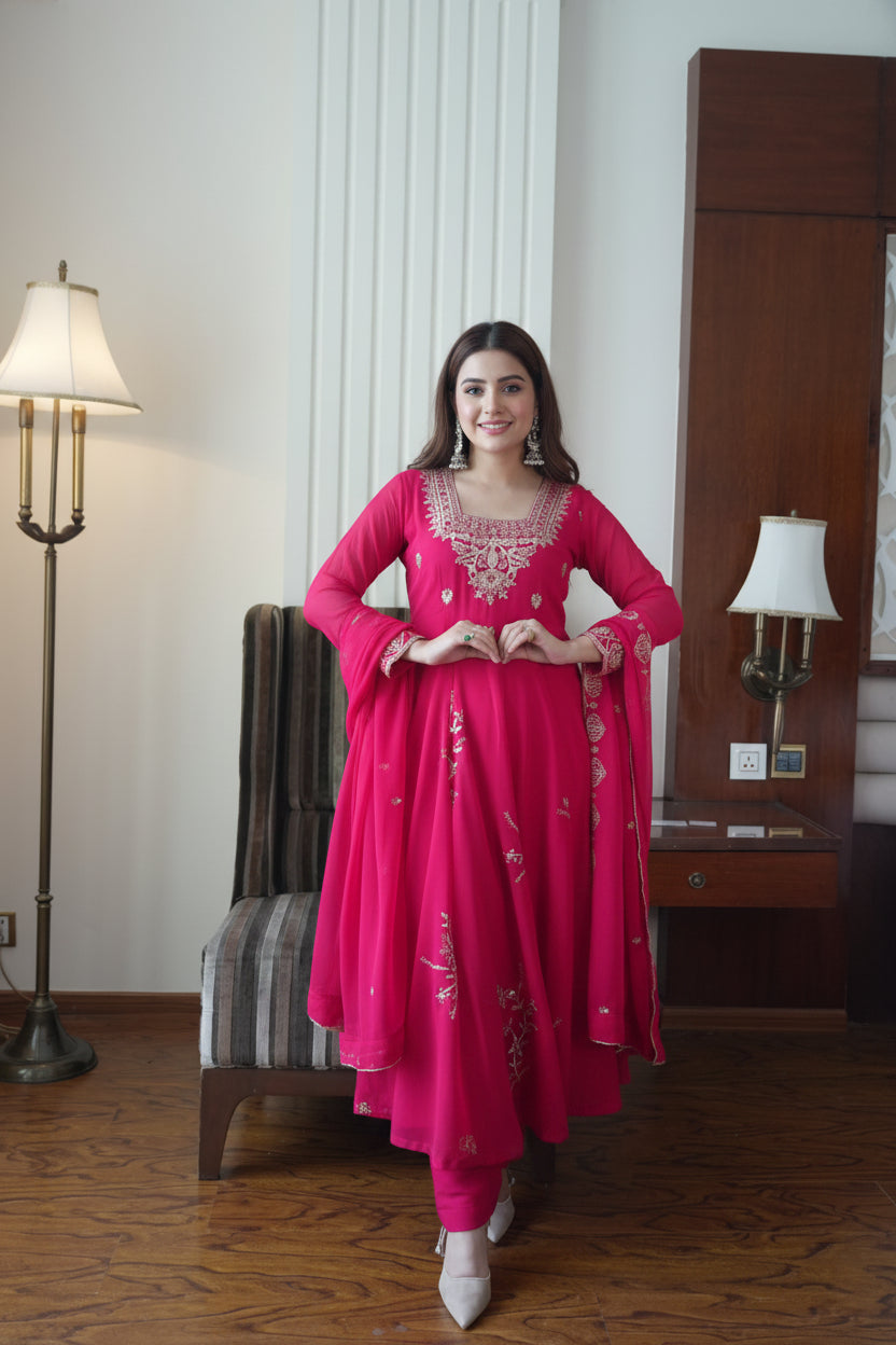 A woman modeling a bright pink traditional Indian outfit with silver embroidery, standing in a furnished room with wooden floors, a lamp, and a chair.